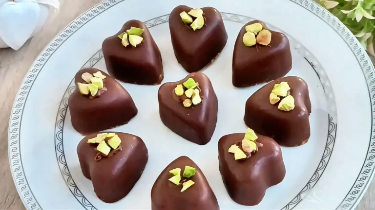 Heart-shaped date pistachio chocolate bites topped with chopped pistachios, served on a decorative plate – healthy no-bake dessert.